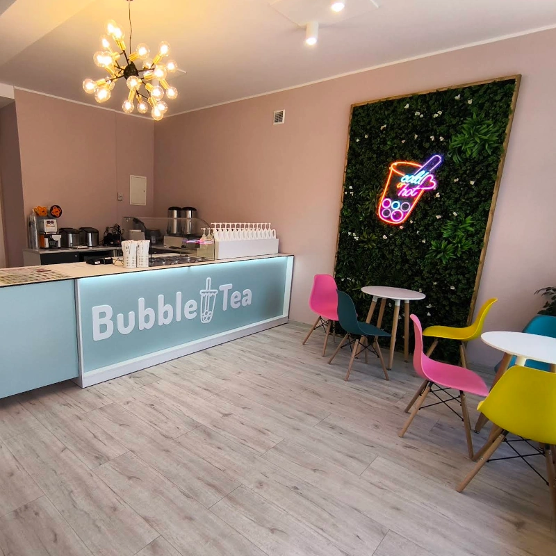 Interior of a Bubble Tea shop featuring a pale blue counter with 'Bubble Tea' text, neon cup sign on a green plant wall, and colorful chairs around small white tables.",