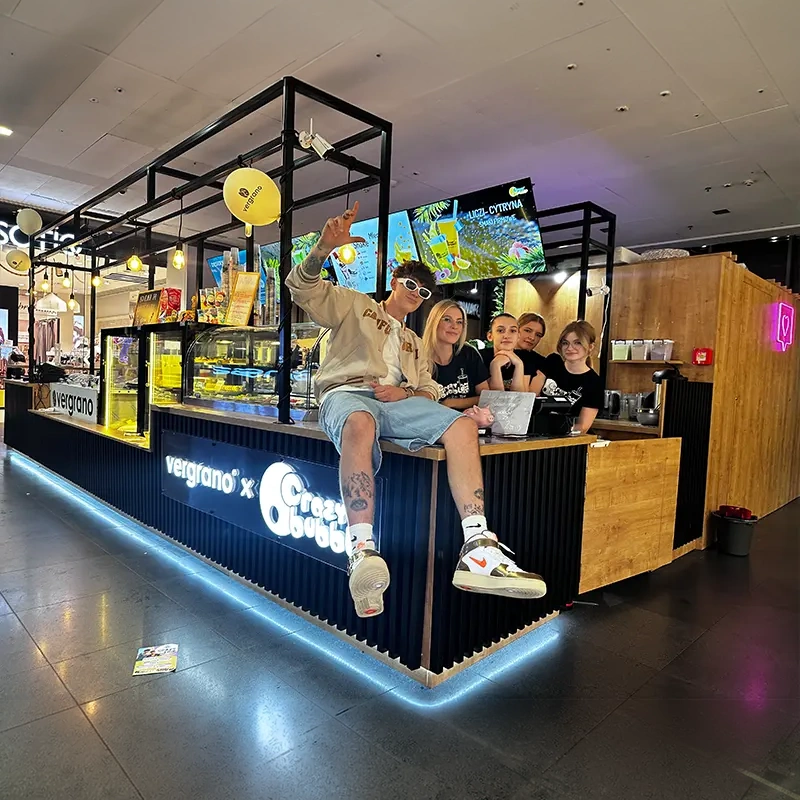Five friends sit on a mall beverage counter, posing for a photo with bright neon signage and drinks behind them in a modern bar area.