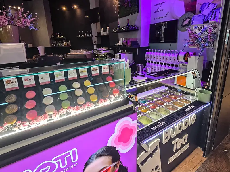 Glass display case with colorful macarons and candy toppings at a bubble tea shop, illuminated by purple lighting outside the counter area.