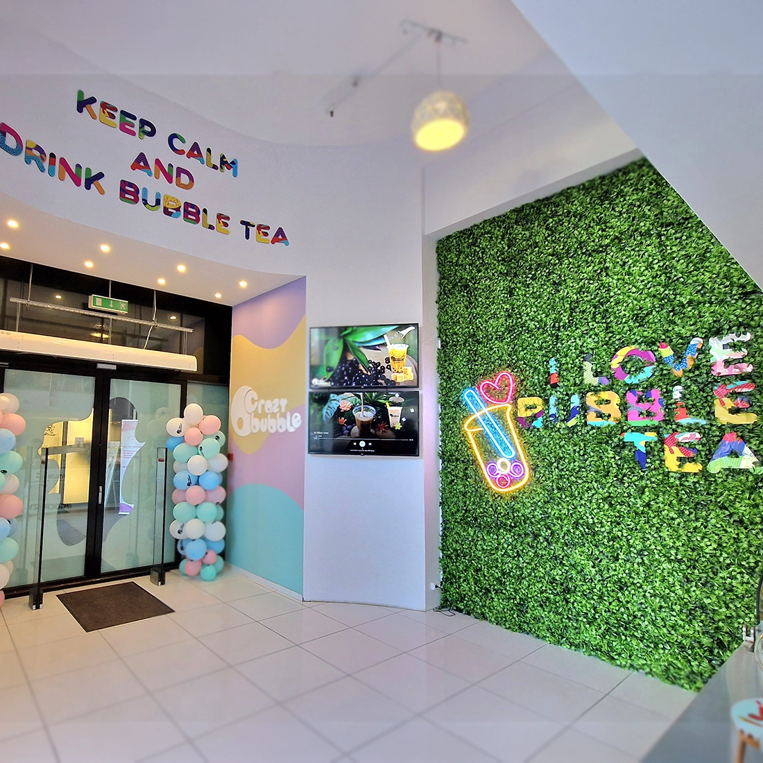Entrance area of a bubble tea shop with pastel balloon columns, glass doors, and a green faux-plant wall with a neon cup sign and colorful 'LOVE BUBBLE TEA' lettering.