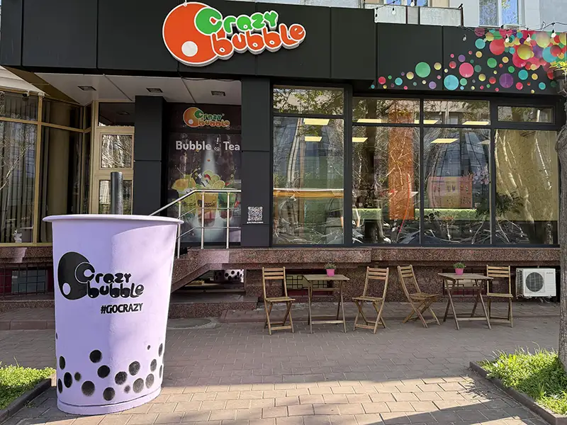 Exterior of Crazy Bubble bubble tea shop with a large purple bubble tea cup prop in the foreground, outdoor tables and chairs, and glass storefront with colorful signages.
