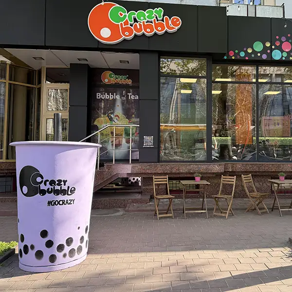 Storefront of Crazy Bubble bubble tea shop with a large purple cup prop in the foreground and outdoor seating along the glass wall.