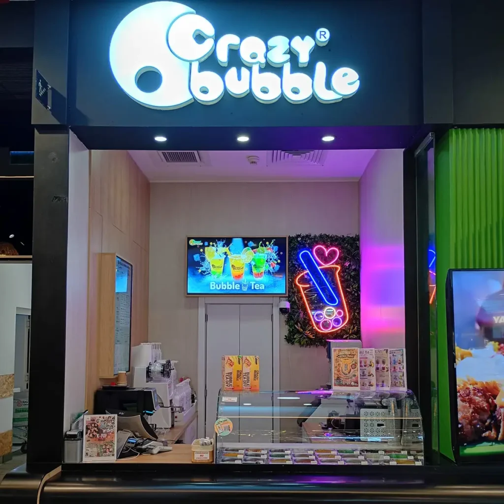 Storefront sign reads 'Crazy Bubble' above a bubble tea shop counter with neon cup sign and menu boards inside.