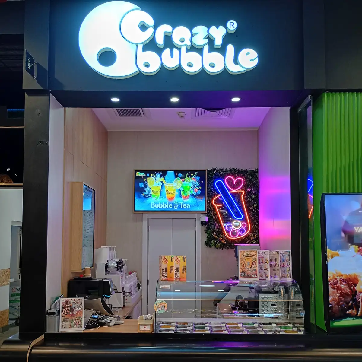 Storefront sign reads 'Crazy Bubble' above a bubble tea shop counter with neon cup sign and menu boards inside.