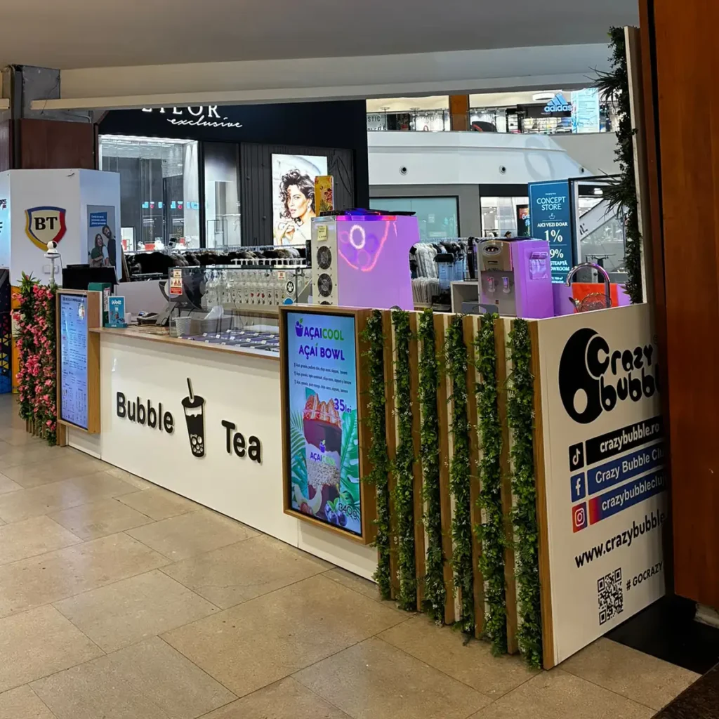 Bubble Tea kiosk in a mall with a white counter showing 'Bubble Tea' and a cup icon, framed by vertical green plants.