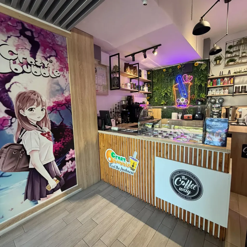 Cafe counter in a modern shop with a neon cup sign, glass display case, and wooden slat front; anime-style poster on the left wall and shelves with equipment behind the counter.