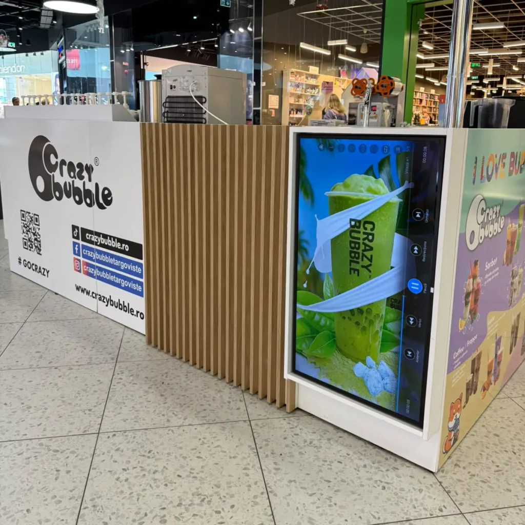 Kiosk in a shopping mall promoting Crazy Bubble with a digital screen showing a green smoothie cup wrapped in white bands and wooden slats around it
