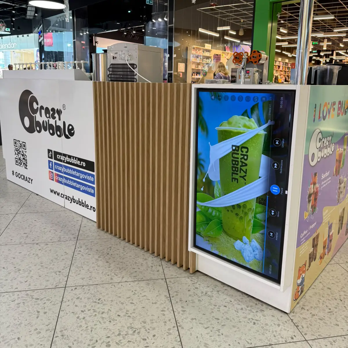 Kiosk in a shopping mall promoting Crazy Bubble with a digital screen showing a green smoothie cup wrapped in white bands and wooden slats around it
