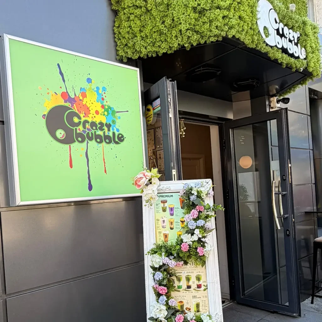 Exterior view of Crazy Bubble cafe: bright green sign with colorful splash logo, open glass door, and a flower-decorated menu board outside.