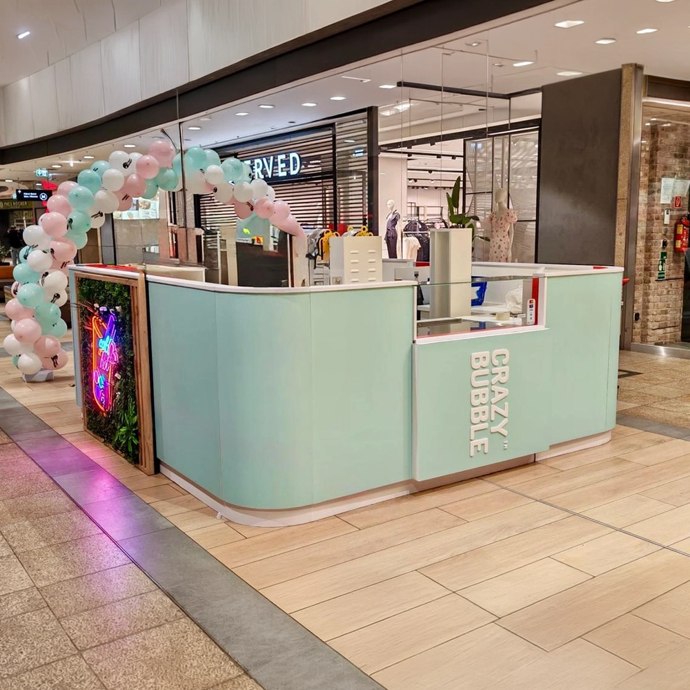Bubble tea kiosk in a mall with a pastel teal counter and a pastel balloon arch, branded 'Crazy Bubble'. Fun, festive storefront.
