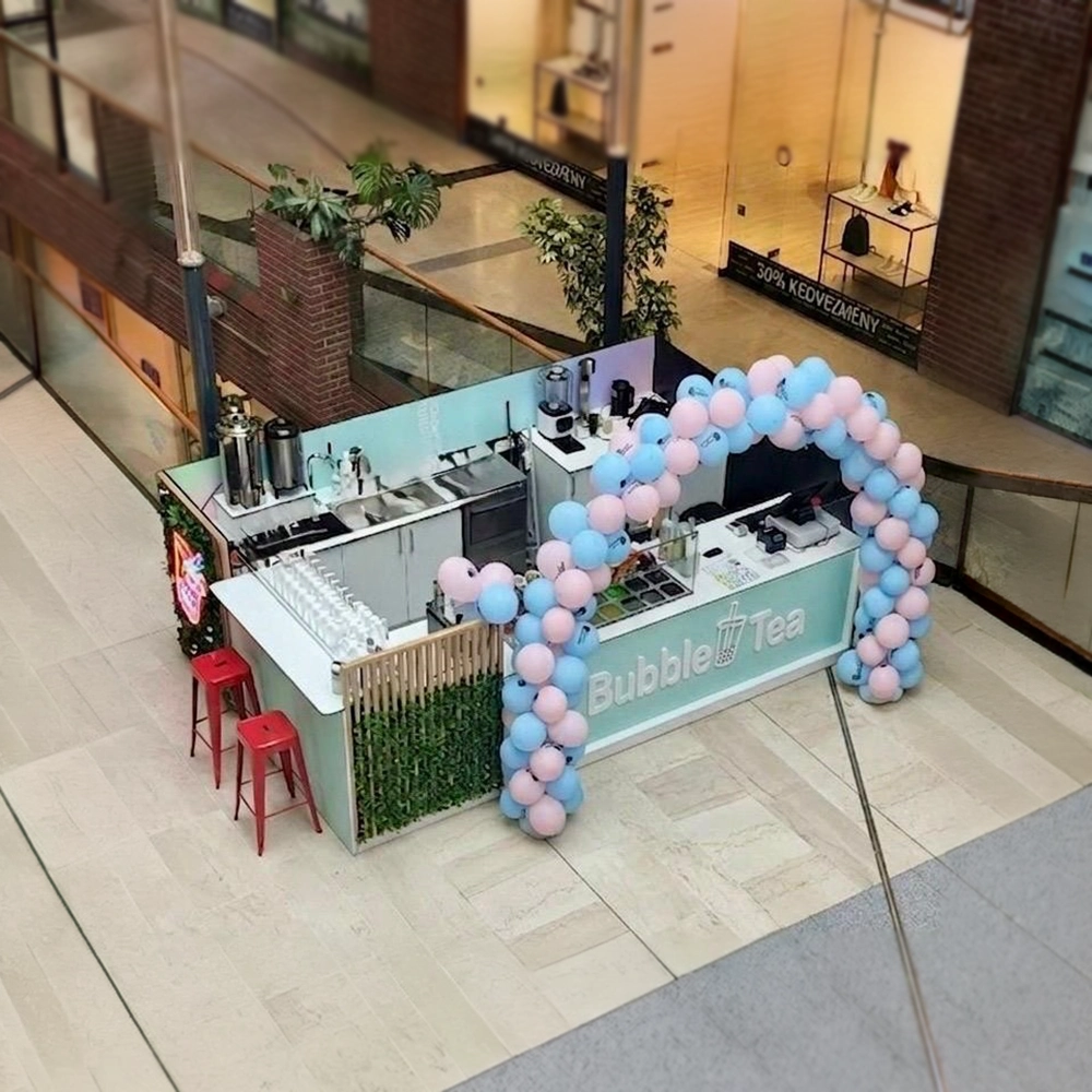 Bubble Tea kiosk in a mall with a pastel blue sign, pastel balloon arch, and red stools at the counter.