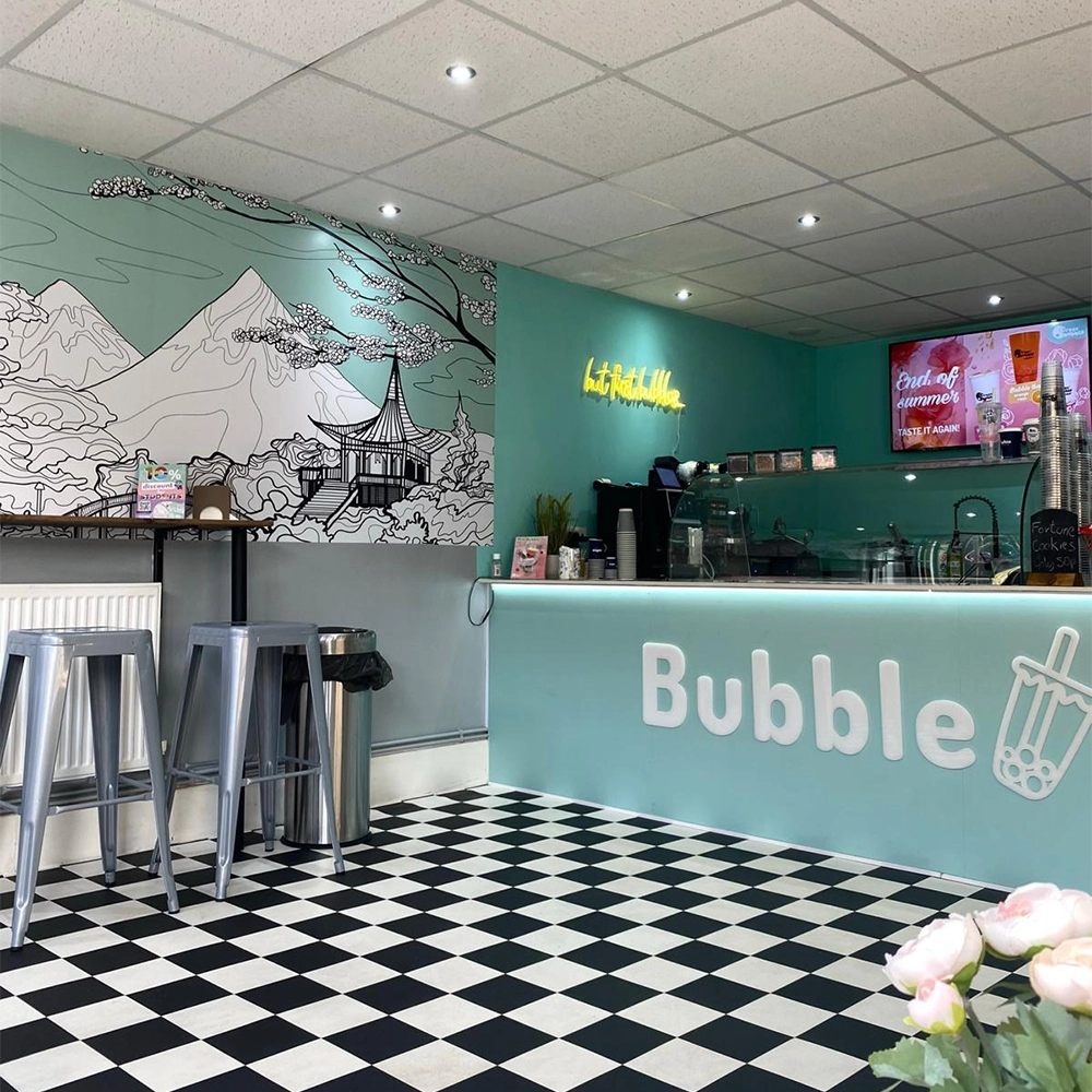 Bright teal ice-cream shop interior with a counter labeled 'Bubble', checkered black-and-white floor, metal stools, mural of mountains and a pagoda, and a neon sign on the wall.