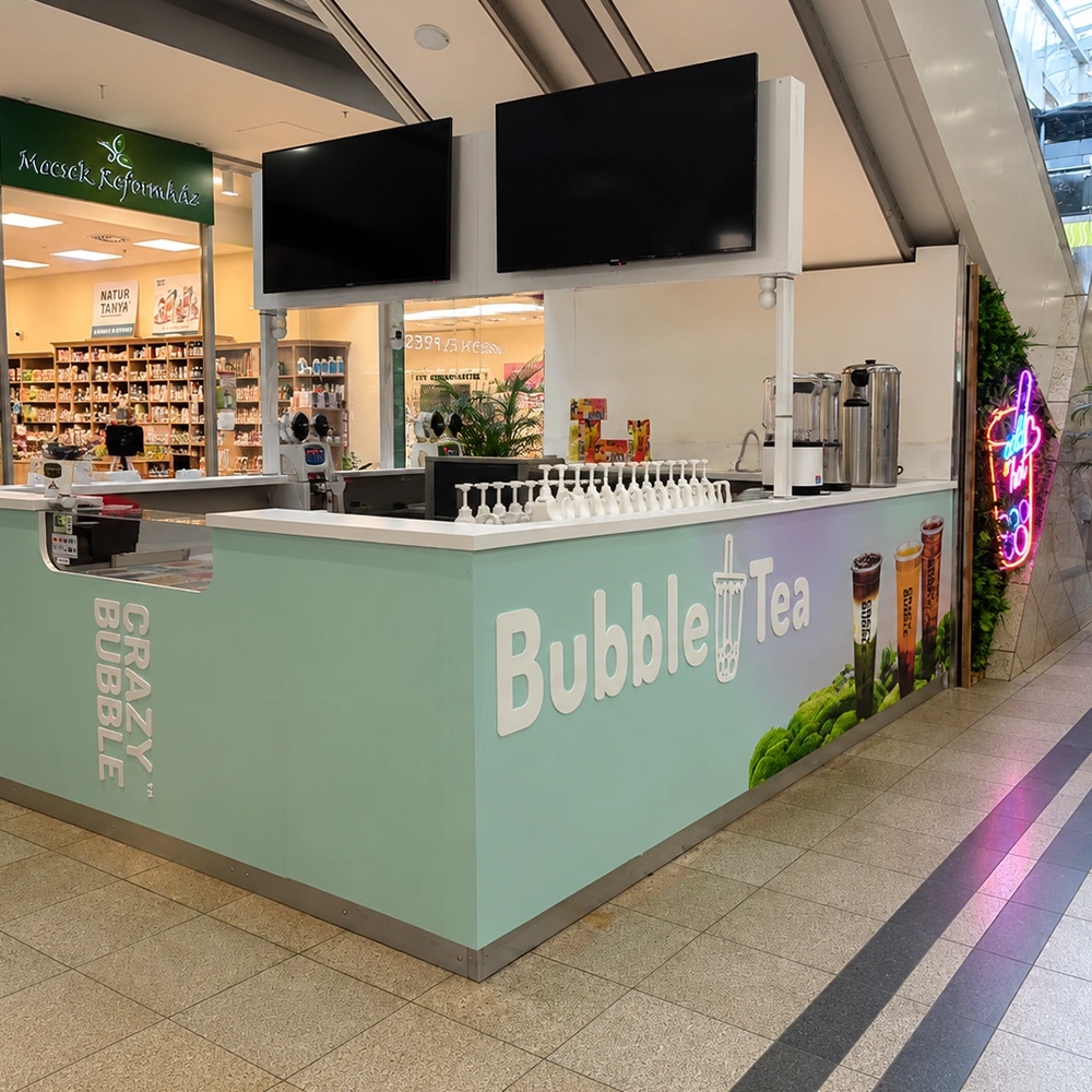 Mint-green bubble tea kiosk in a mall with cups lined up, beverage dispensers, and a large 'Bubble Tea' sign above; two flat-screen monitors hang overhead.
