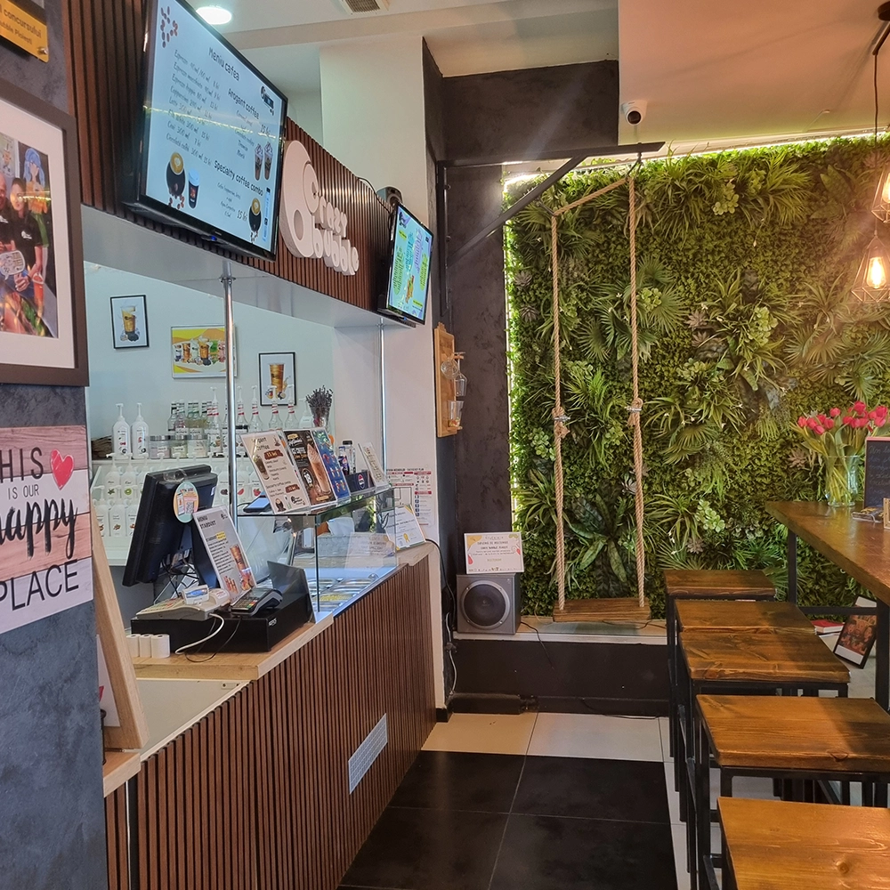 Cafe interior with a long wooden counter, glass display case, and digital menu boards above. A vertical garden wall with hanging ropes is to the right; wooden stools line a table area.