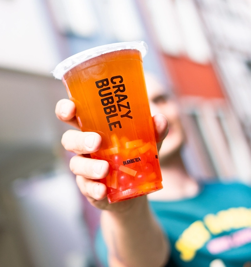 Hand holding an orange plastic cup labeled 'CRAZY BUBBLE' with a white lid, containing an orange beverage with candy pieces visible inside.