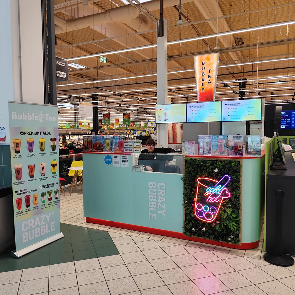 Bubble tea kiosk in a mall-style food court with a turquoise counter, neon cup sign, and menu screens overhead.