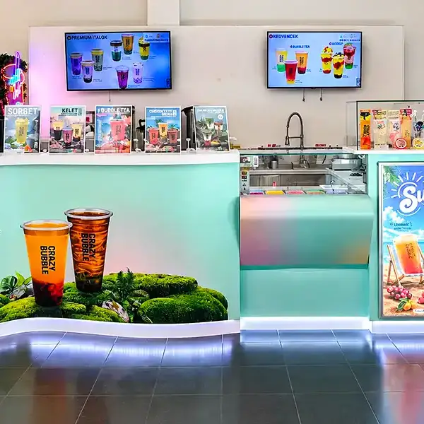 Bright beverage kiosk with teal counter, two large iced drinks on a mossy display, and digital menu boards above the counter selling bubble tea flavors.