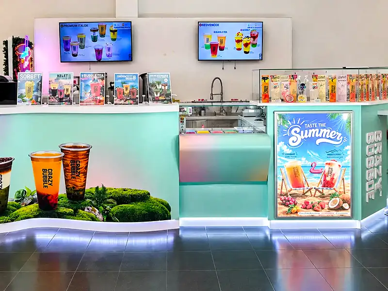 Mint-green beverage counter with digital menus, cup displays, and a 'Taste the Summer' fridge poster on the right.