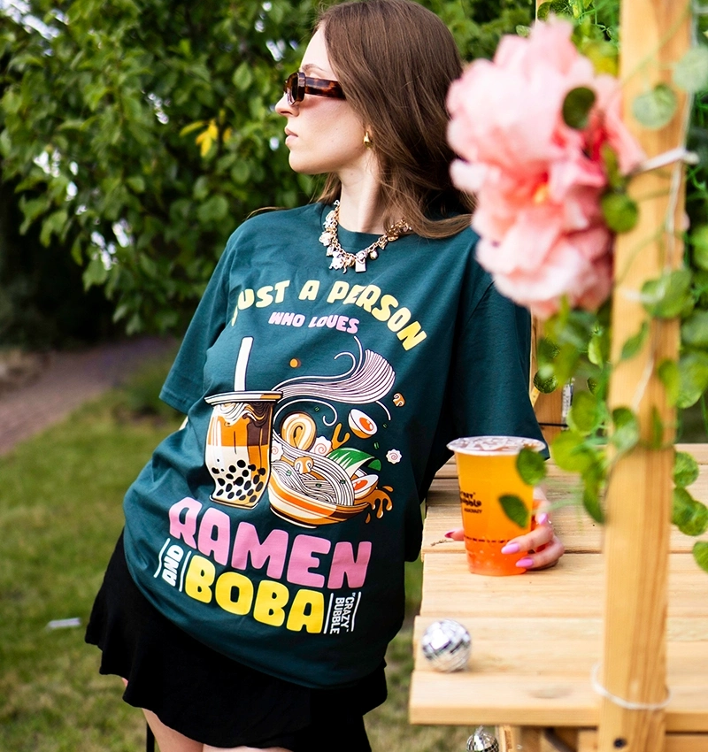 Woman in sunglasses leaning on a wooden railing, wearing a teal graphic t-shirt about ramen and boba, holding an orange drink outside.