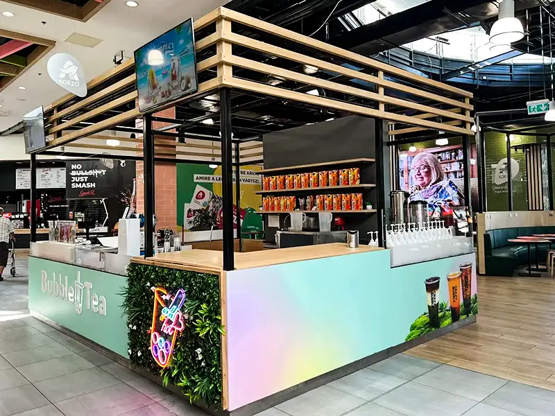 Kiosk-style bubble tea stand in a mall with a pastel gradient counter wrap, wooden frame canopy, and shelves of colorful cups behind the counter.