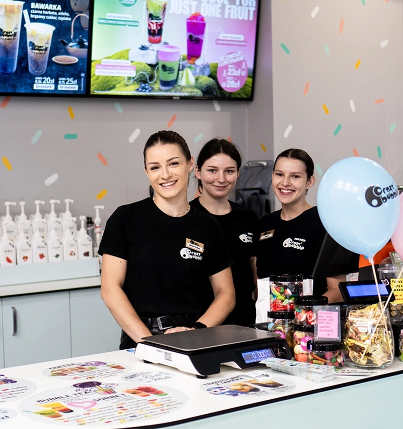 Three smiling staff members in black uniforms stand behind a smoothie shop counter with balloons and colorful drink posters on the wall.