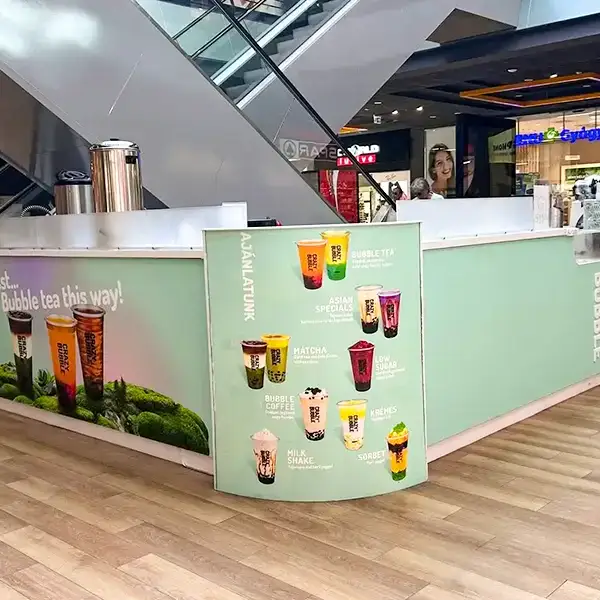 Mall beverage kiosk with a curved menu displaying colorful bubble tea drinks; escalator and shops in the background, mall setting.