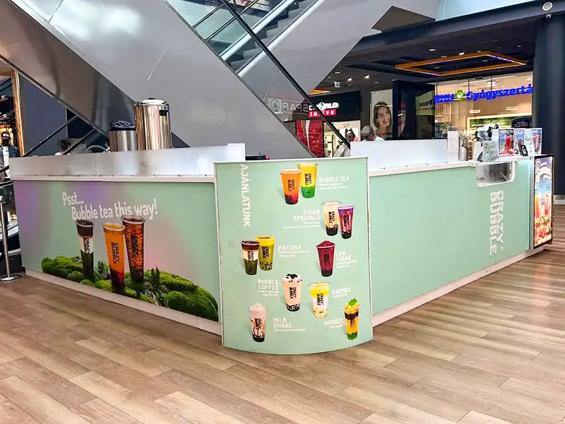 A bubble tea kiosk in a mall with pastel-green panels, menu cups displayed on a curved stand, and an escalator in the background.