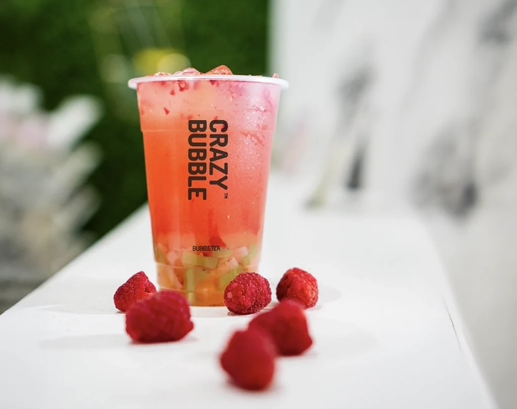 Pink fruit bubble tea in a clear cup with the Crazy Bubble logo, on a white table with fresh raspberries scattered around.