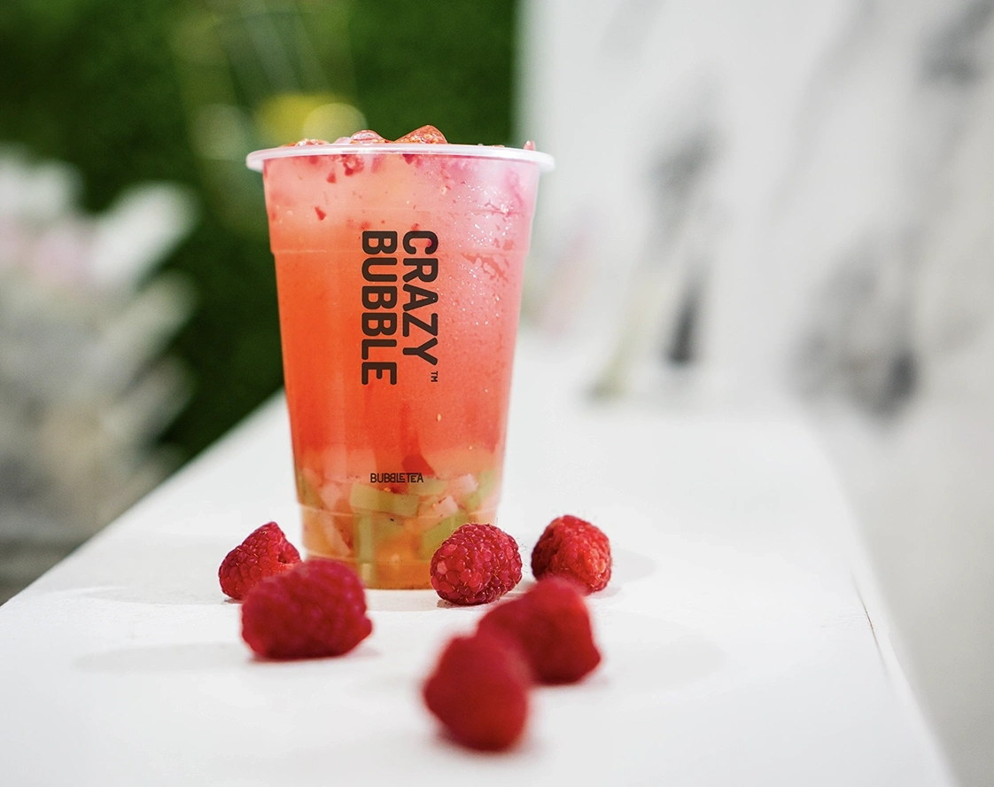 Pink fruit bubble tea in a clear cup with the Crazy Bubble logo, on a white table with fresh raspberries scattered around.