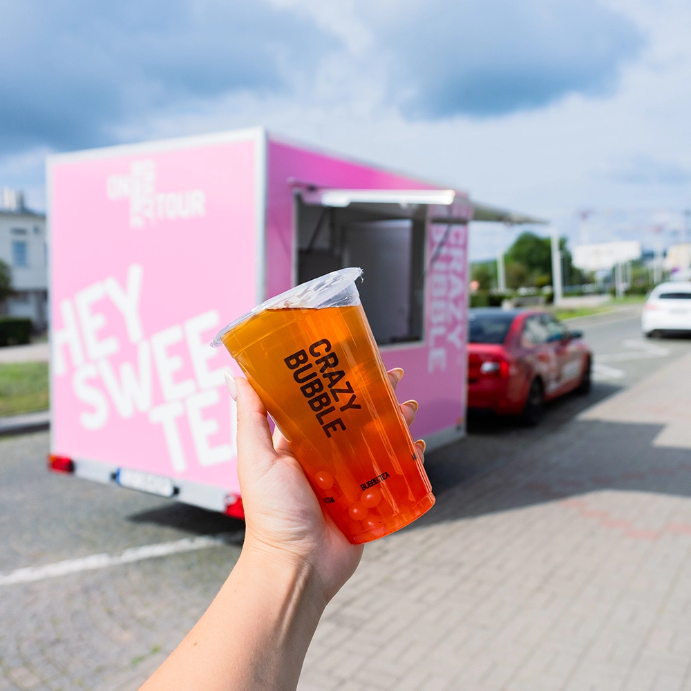 Close-up of a hand holding an orange bubble tea cup labeled 'Crazy Bubble' with a pink beverage trailer in the background.