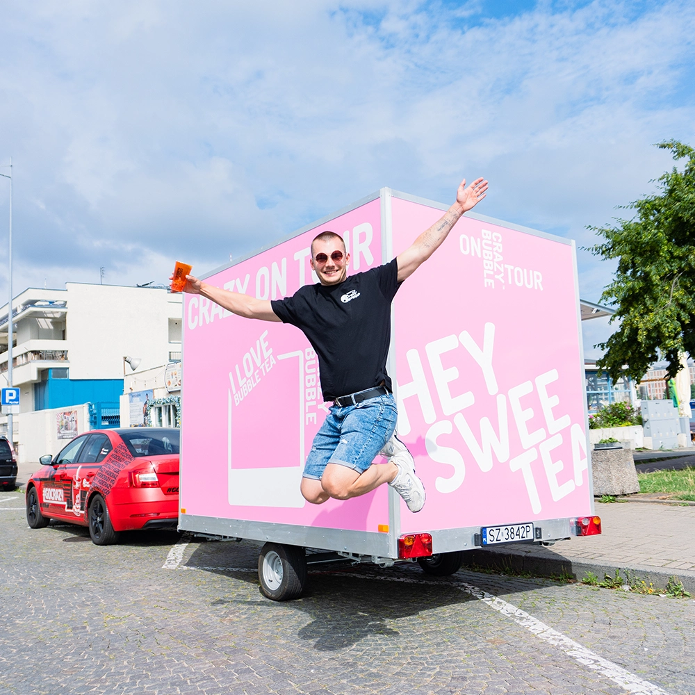 Man in a black t-shirt and denim shorts jumps with arms open beside a bright pink promotional trailer. He holds an orange cup; a red car is in the background.