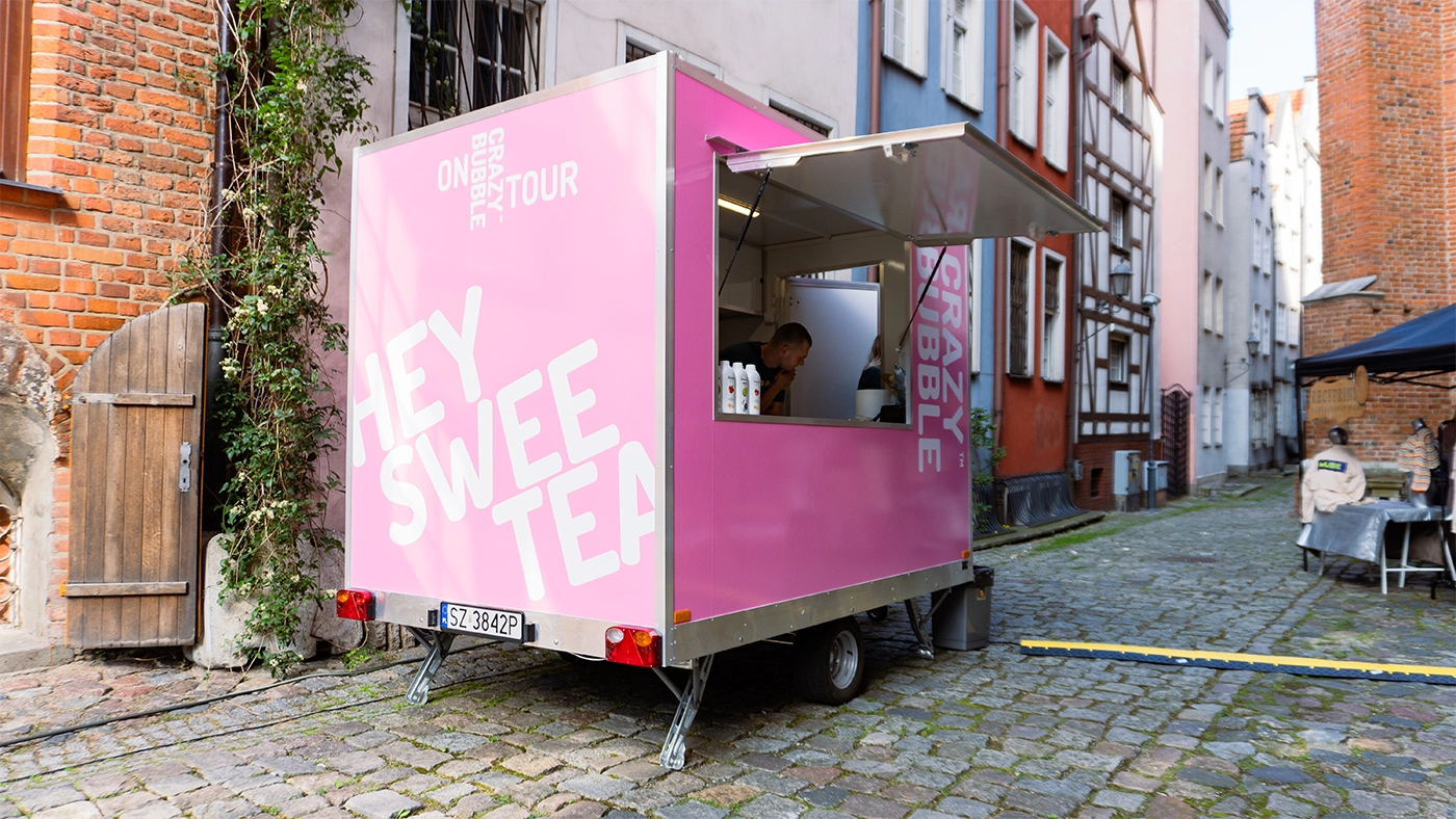 Pink mobile tea trailer on a narrow cobblestone street, serving beverages from an open window.