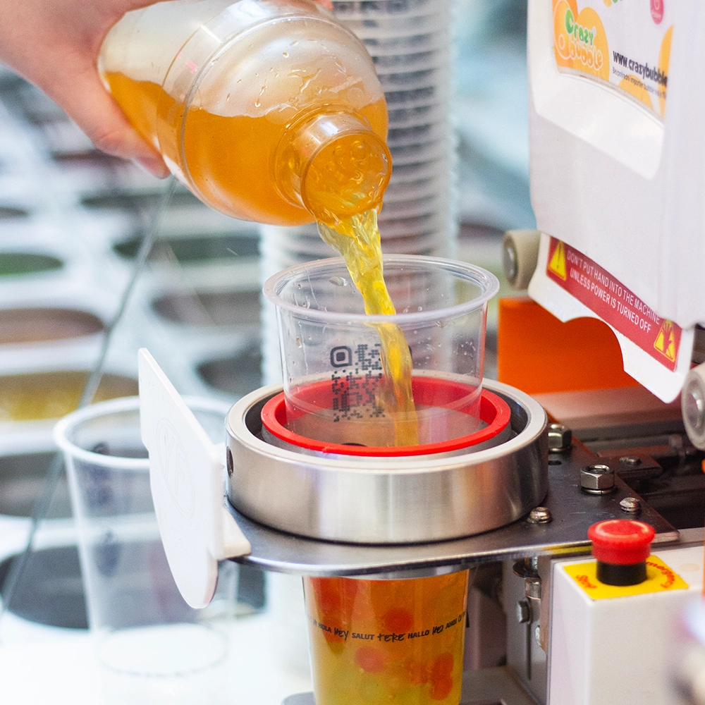 Close-up of orange juice being poured from a bottle into a clear cup under a stainless steel beverage dispenser.