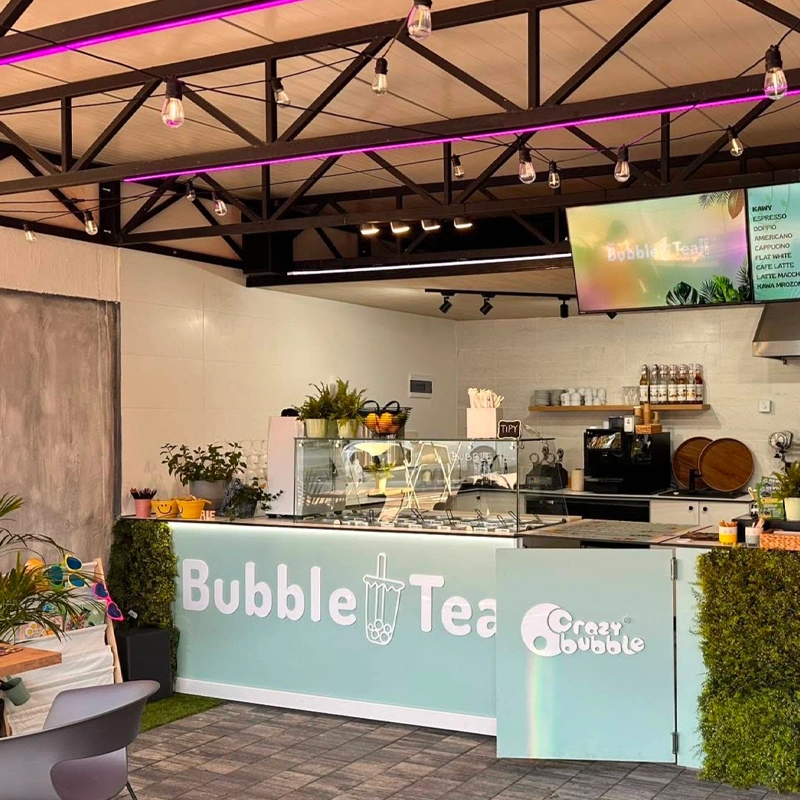 Bubble Tea shop counter with a pastel blue front, glass display case, and potted plants under purple string lights.