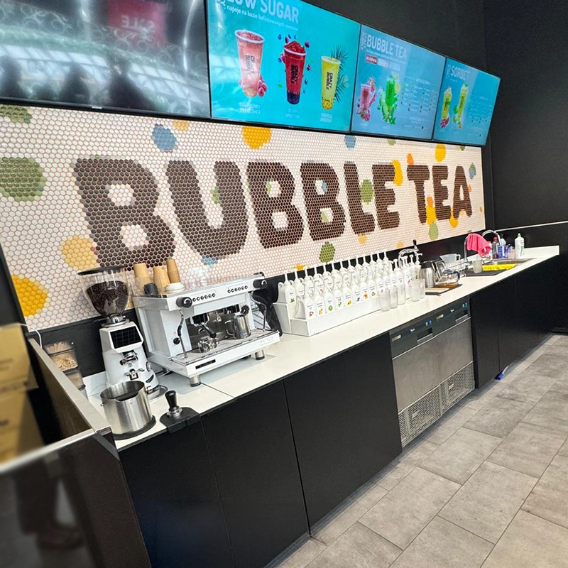 Bubble tea shop counter with an espresso machine, syrups, cups, and a colorful mosaic wall reading 'BUBBLE TEA' and digital menu panels above.