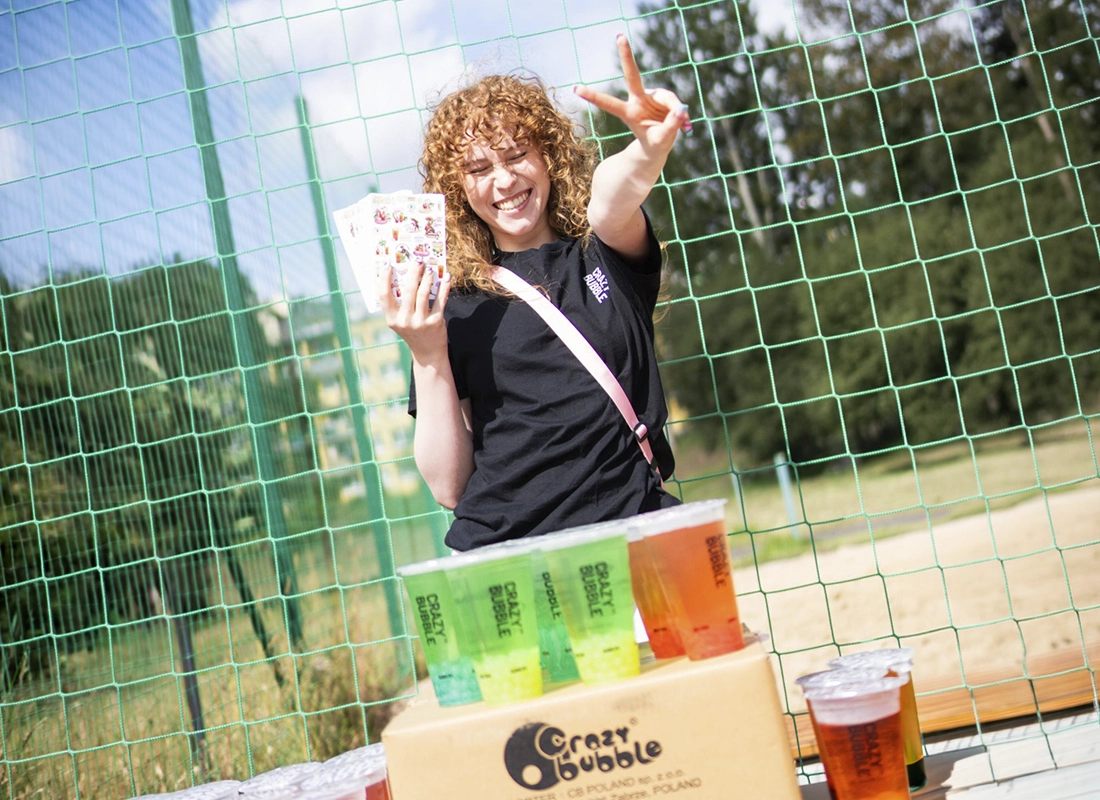 Smiling woman with curly hair holds cards and shows a peace sign beside a table of colorful beer cups outdoors behind a green fence.