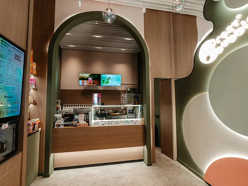 Cafe counter in a contemporary shop with a curved green archway, glass pastry display case, and digital menu screens in the back area.